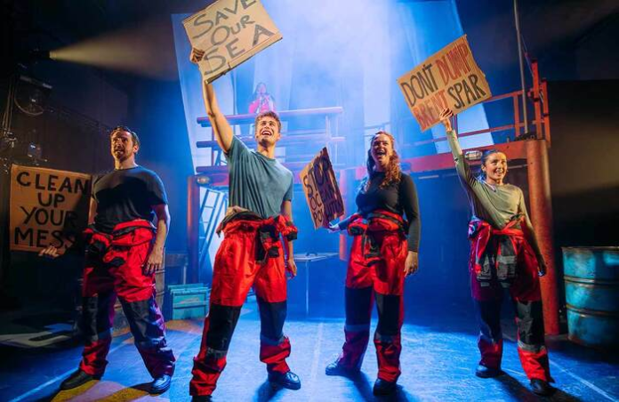 Four actors dressed in red boiler-suits, stand on stage as environmental activists, with an oil rig behind them, holding up signs saying 