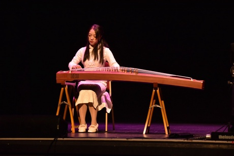 A woman sits on stage with a large guzheng: a traditional Chinese zither.