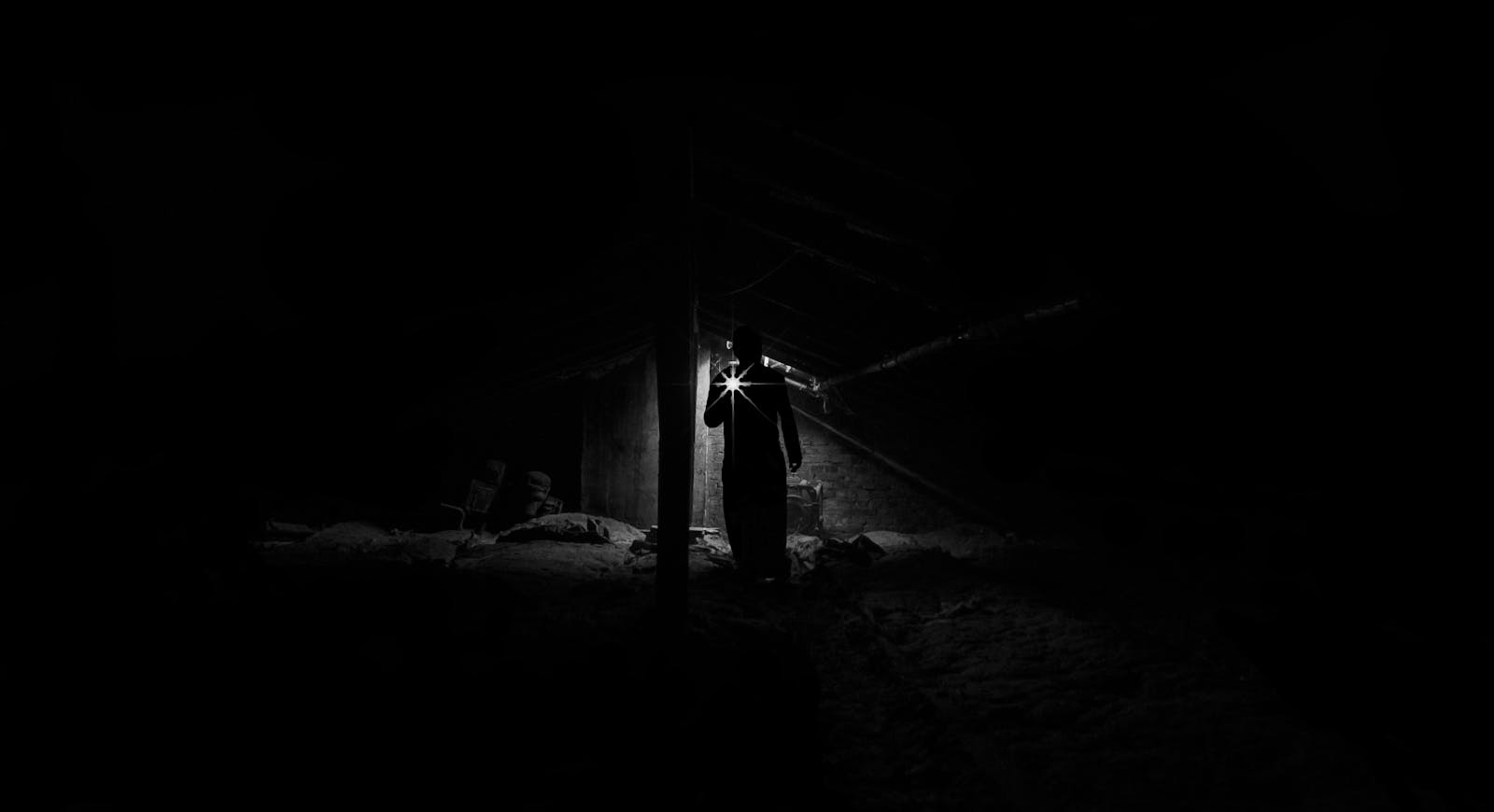 A person in the dark with a flashlight. A shadowy figure explores a dimly lit, eerie attic with a flashlight.