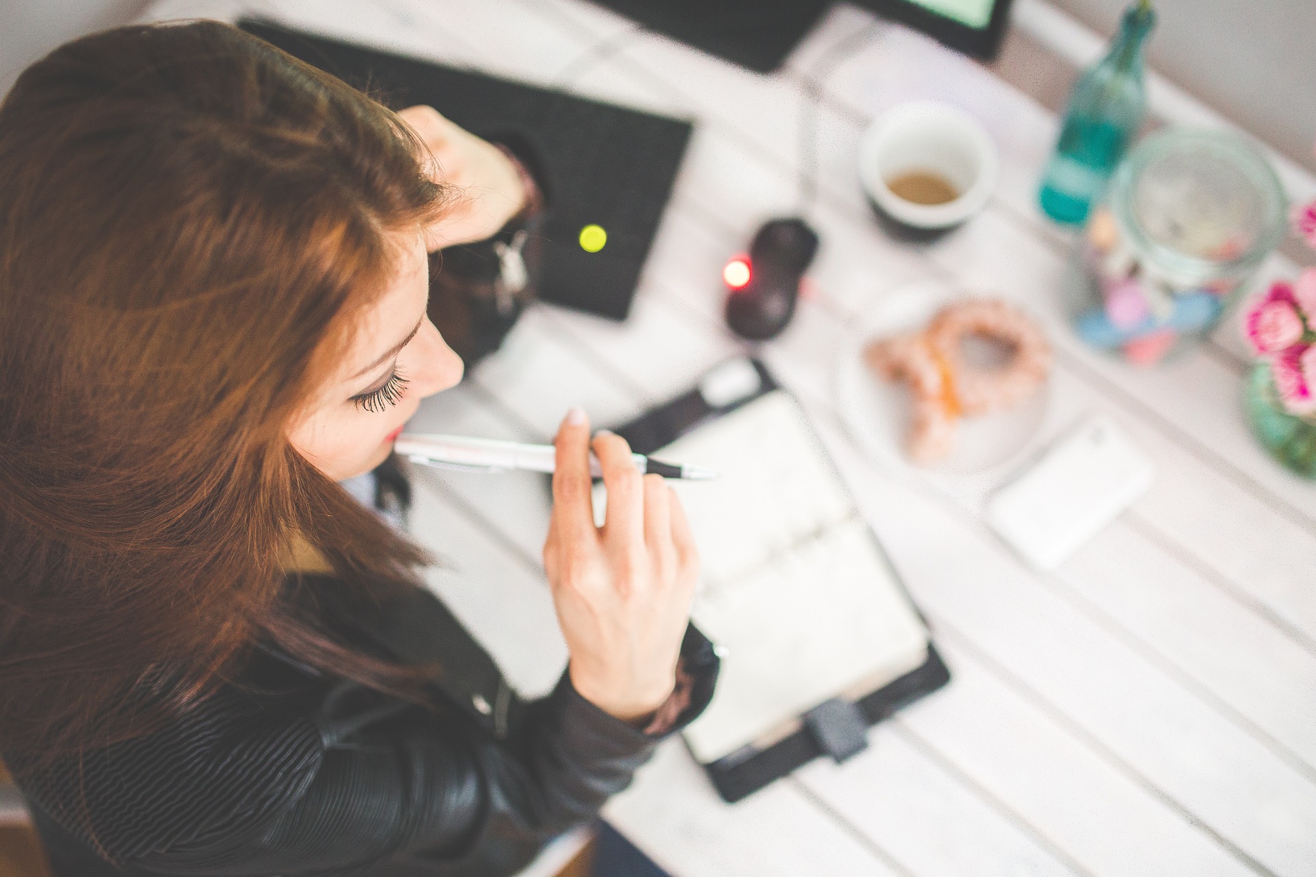 A woman studying. A female student with brown hair, wearing a leather jacket sat at a desk with a pen in her mouth. There is a notebook, keyboard, mouse, coffee and a doughnut on the desk.
