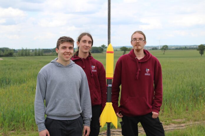 A photo of the University's Rocketry Society, 3 members stand in frame with a yellow and black rocket.