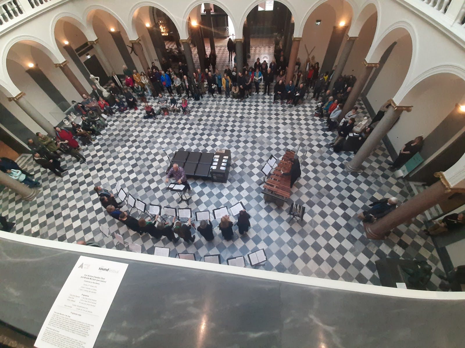 Con Amina Chamber Choir and Brodie McCash. A wide-angle photo taken from a balcony, overlooking a choir in the centre of the floor with a conductor and piano, surrounded by a modest crowd.