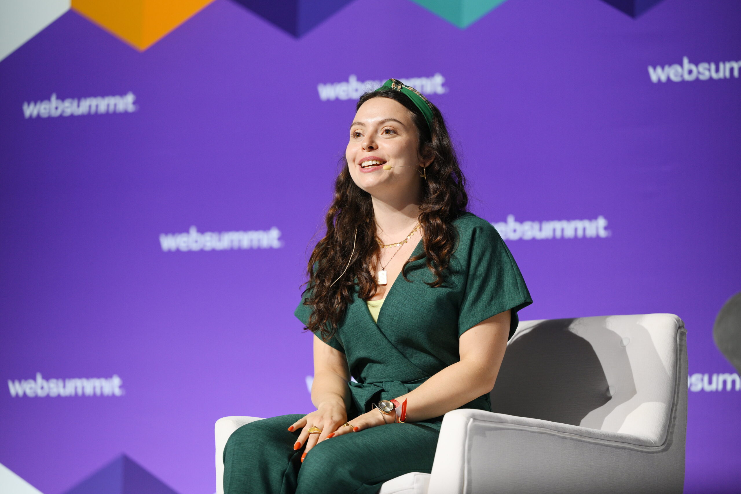 A photo of Sophia Smith Galer on stage at Web Summit, 2022. A photo of Sophia Smith Galer on stage at Web Summit, 2022. She is sitting on a cream sofa chair, wearing an emerald green jumpsuit.