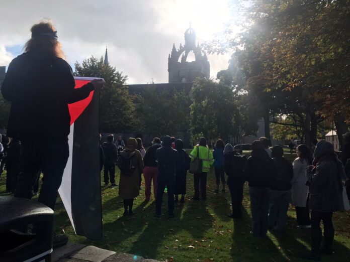 A group of people gathered for a demonstration at University of Aberdeen. A person is holding a Palestine flag.