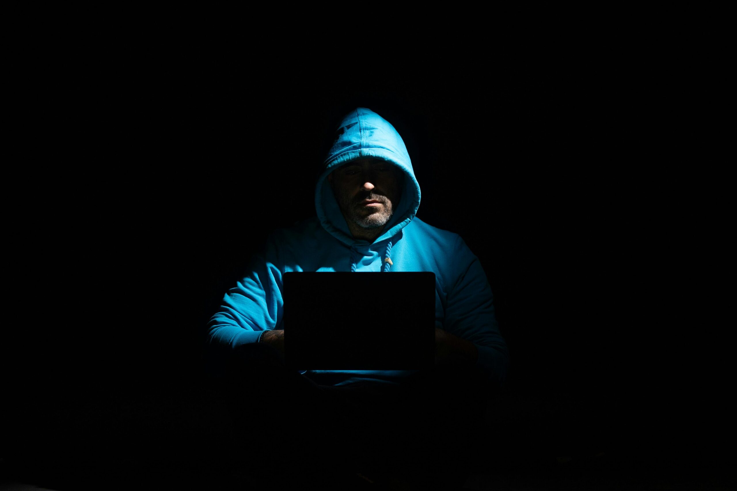 The internet is not a safe place… or is it? Person in blue hoodie sitting in the dark with a laptop.